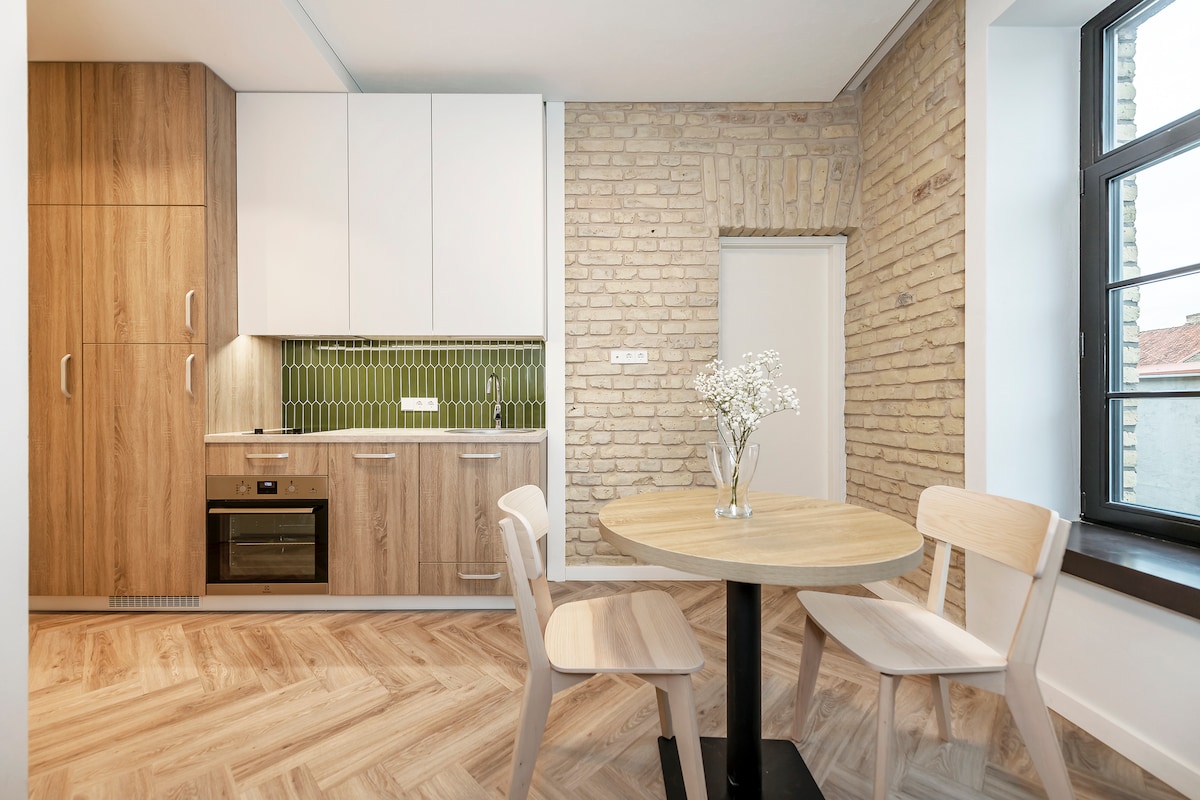 A modern kitchen area showcases light wood cabinetry harmonized with a textured brick wall. A round table with two wooden chairs sits in front of a window, allowing natural light to fill the space. Green tile accents provide a fresh touch to the design.