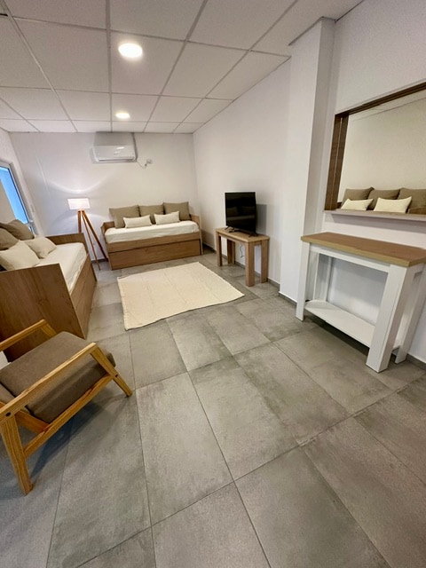 A bright and airy room features a spacious sofa and a daybed with soft, neutral bedding. A small wooden table and chair provide a seating area, while a mirror adds depth. Natural light floods in from a window, enhancing the minimalist decor.