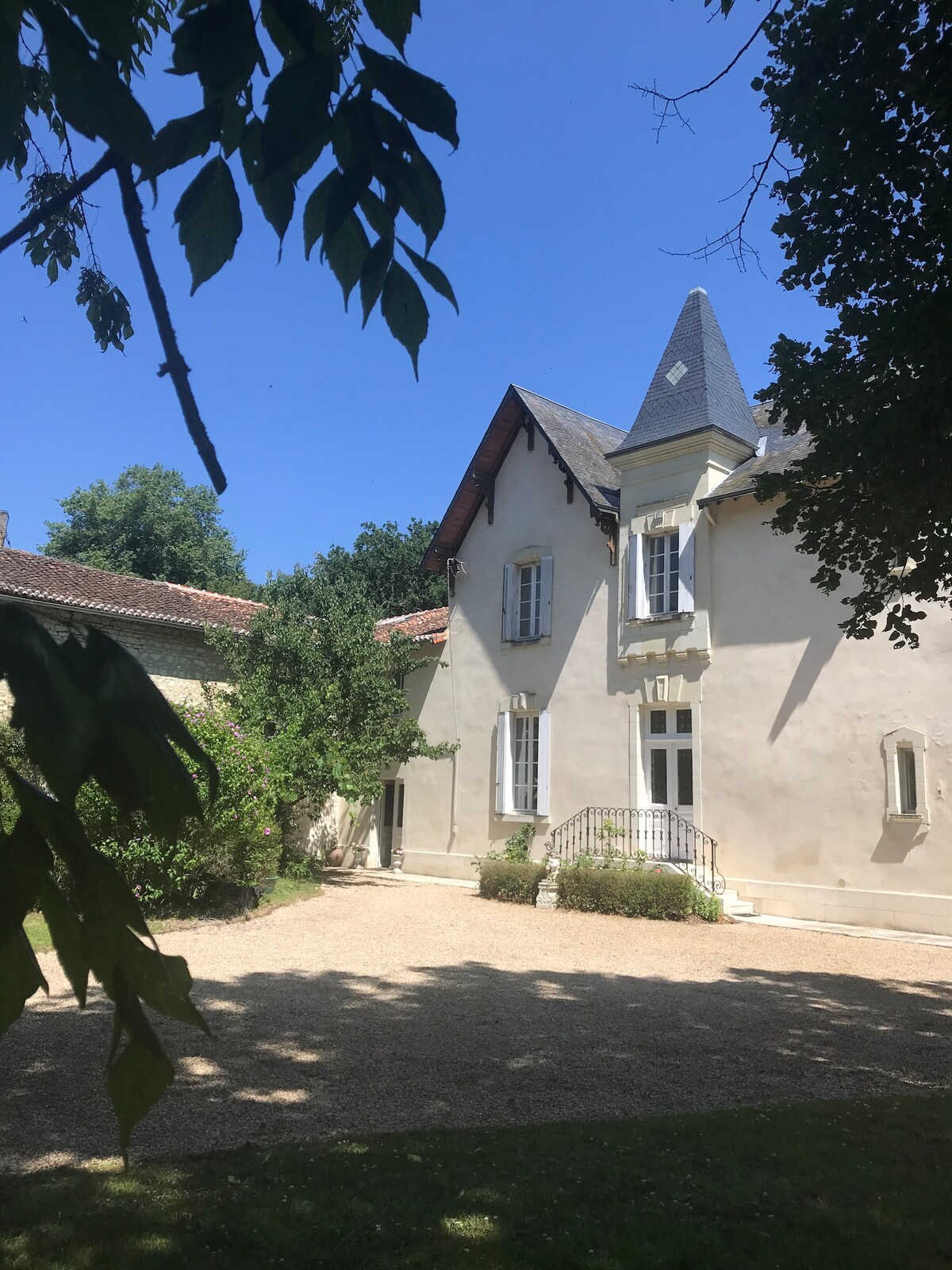A charming manor exterior is framed by lush greenery, showcasing a combination of stone and decorative architectural elements. Sunlight filters through the leaves, highlighting a pebbled pathway leading to an inviting front entrance with elegant windows and a gabled roof.