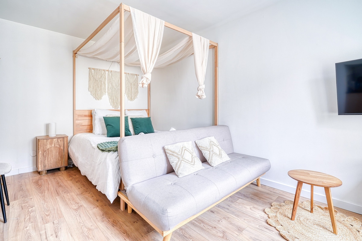 A spacious room features a wooden canopy bed adorned with draped fabric and soft pillows. A versatile sofa bed is positioned nearby, complemented by decorative cushions. Light-colored walls enhance the overall brightness, while a flat-screen TV is mounted on an adjacent wall.