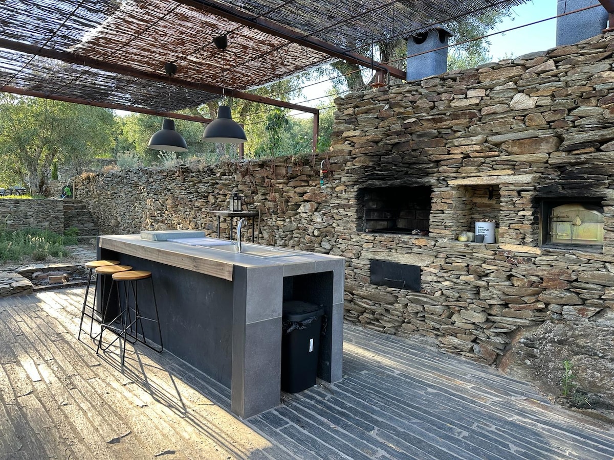 An outdoor kitchen area is highlighted by a large stone countertop and seating with three black bar stools. Natural stone walls frame built-in ovens, while a pergola provides overhead shading. Sunlight filters through, creating a warm and inviting cooking space surrounded by greenery.