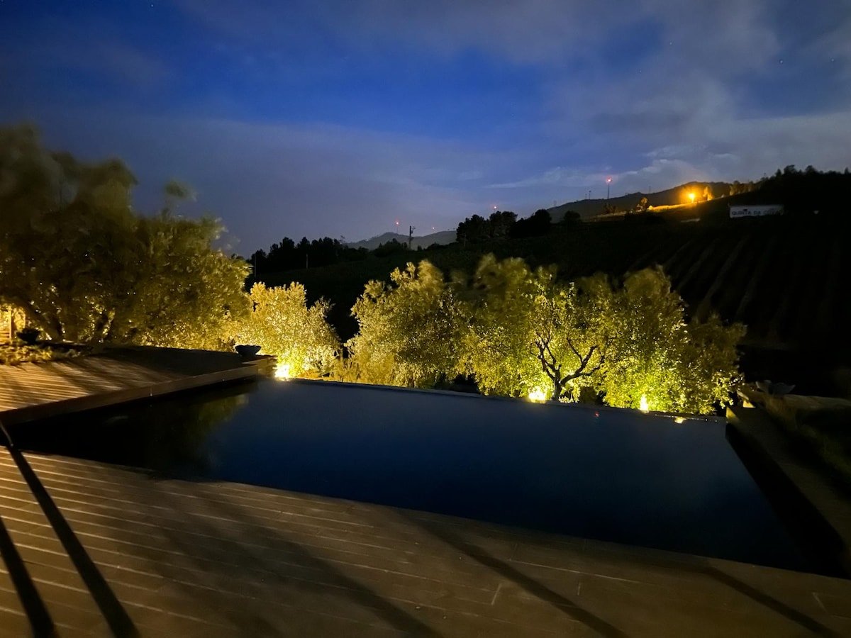A serene outdoor area features an infinity pool reflecting soft night lights, surrounded by lush greenery. The landscape of rolling hills can be seen in the distance, with subtle illumination highlighting the natural features against the evening sky.