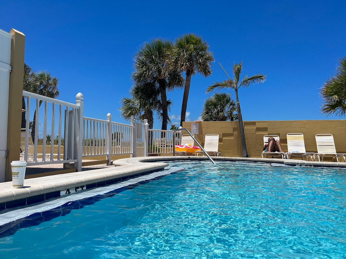 The heated saltwater pool is surrounded by a white fence and features several sun loungers. Palm trees provide a tropical backdrop under a clear blue sky, with a glimpse of the beach visible beyond the pool area.