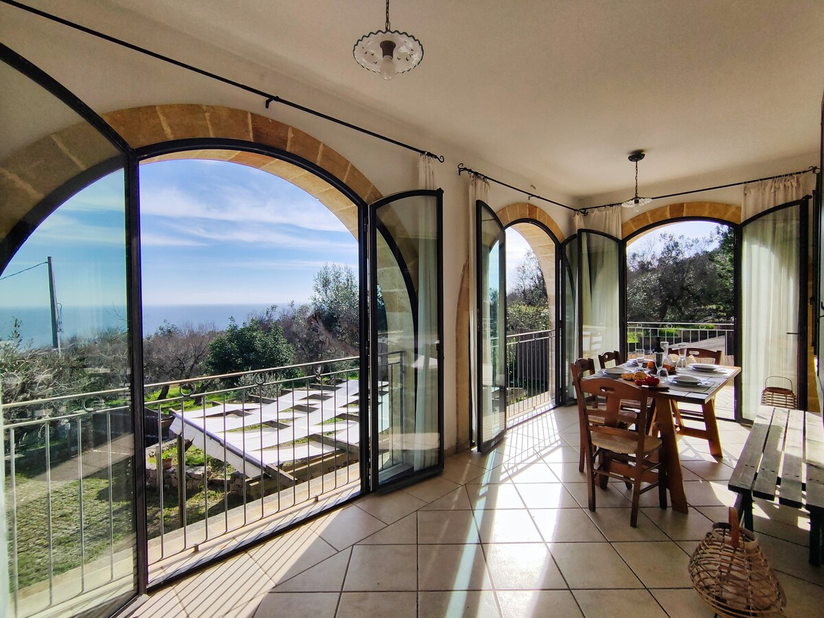 Bright dining area is framed by large arched windows, offering panoramic views of the sea and surrounding greenery. A wooden table is set for meals, surrounded by chairs, while the tiled floor enhances the spacious feel of the room.
