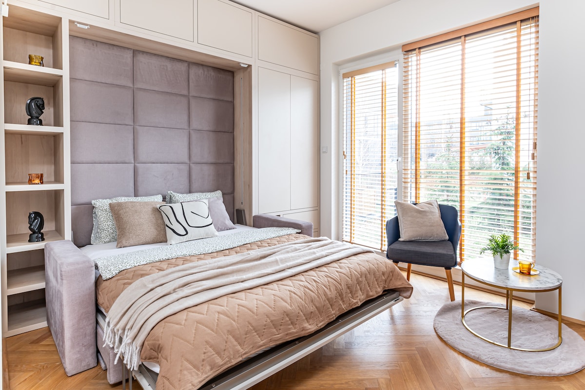 A modern studio features a comfortable fold-out bed with soft bedding, situated against a stylish padded wall. Natural light enters through large windows adorned with wooden blinds, illuminating a seating area with a cozy chair and a small round table.
