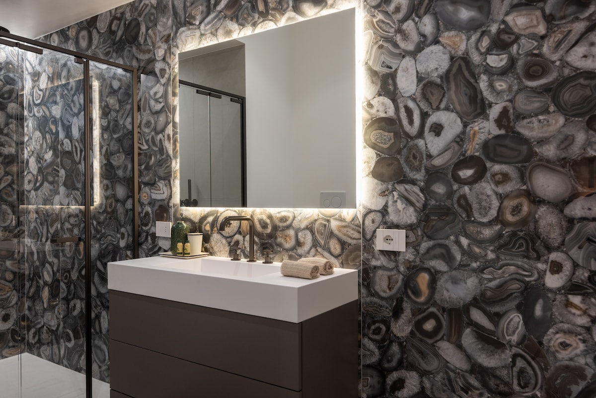 A modern bathroom features an elegant countertop with a stylish basin and dual faucets. The walls are adorned with intricate stone patterns, and soft lighting surrounds a large mirror, enhancing the contemporary feel of the space.
