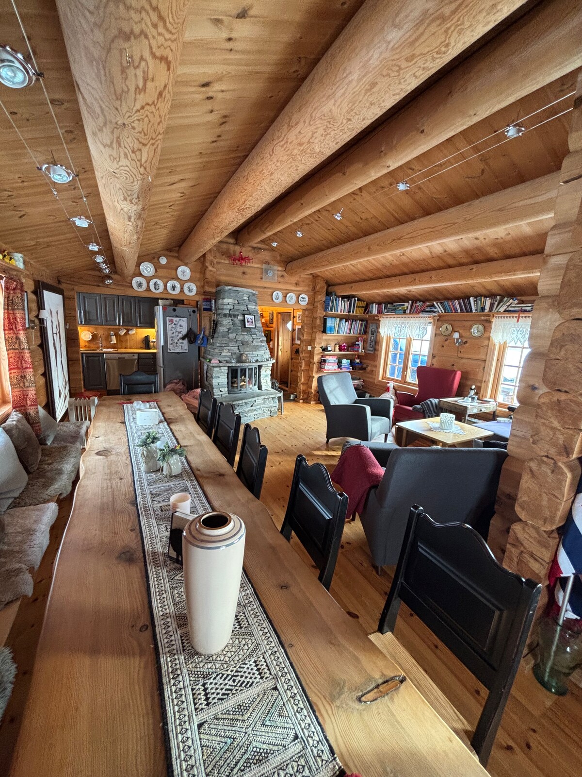 The interior of the timber cabin features a long wooden dining table with dark chairs and a decorative runner. Comfortable seating areas are arranged around a central fireplace, with exposed wooden beams overhead and bookshelves along the wall. Windows allow natural light to brighten the space.