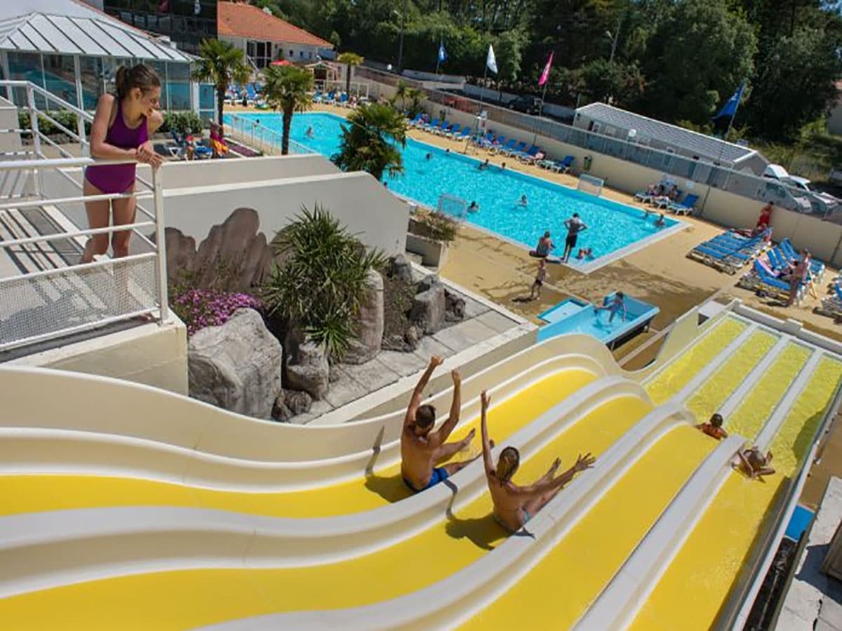A water park area is shown with vibrant yellow water slides descending towards a shallow pool. Sunbathers can be seen relaxing on lounge chairs surrounding the pool, while families enjoy various water activities in the clear blue water. Colorful flags gently wave in the background.