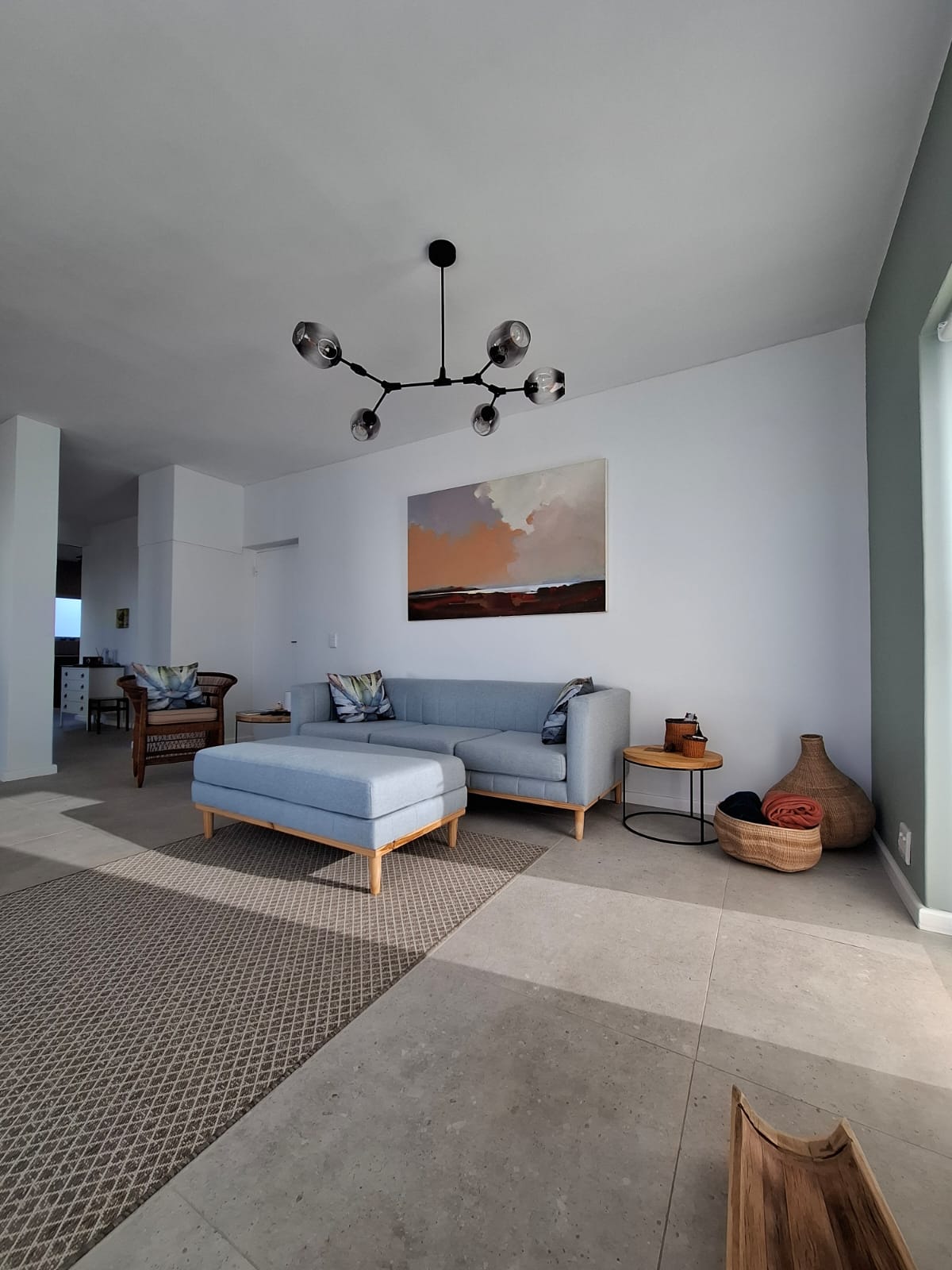 A light and airy living area features a blue sectional sofa paired with a small wooden table. Above, a modern light fixture hangs from the ceiling. The floor is tiled, with a textured area rug adding warmth. A large artwork depicting a cloudy sky is mounted on the wall.