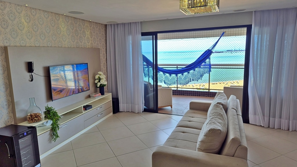 A comfortable living area is presented, featuring a spacious sofa facing a mounted television. Large windows offer a view of the ocean, while a hammock hangs on the balcony. Natural light filters through sheer curtains, creating a relaxed atmosphere.
