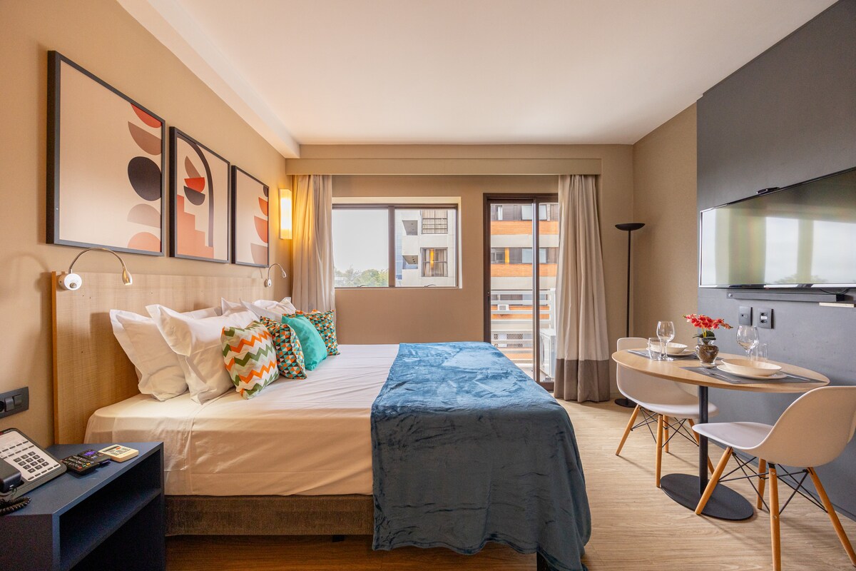 A modern studio features a spacious bed adorned with colorful throw pillows. A dining area is set for two, complemented by a sleek flat-screen TV mounted on the wall. Natural light filters through a large window, highlighting the contemporary artwork above the bed.