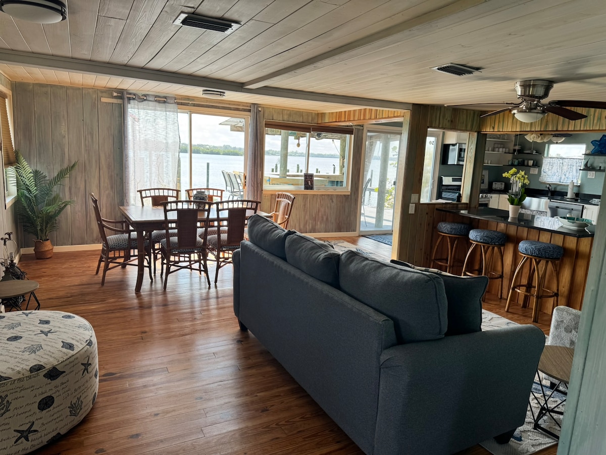 A cozy living area showcases a spacious gray sofa facing large windows with views of the water. A dining table with six wooden chairs is positioned nearby, while a well-equipped kitchen is visible in the background, featuring wooden cabinetry and bar seating.