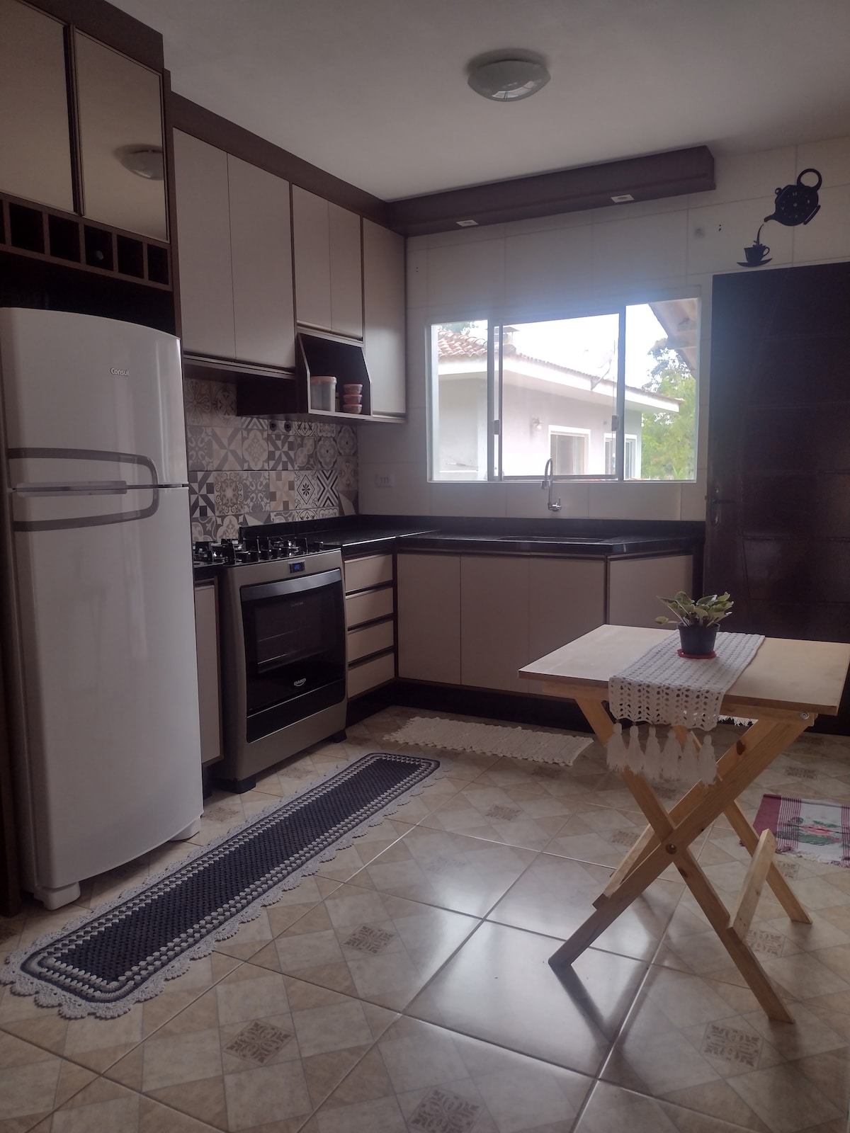 The kitchen features a combination of cabinets and modern appliances, including a white refrigerator and a gas stove. A small wooden table sits on patterned floor tiles, next to a window allowing natural light to brighten the space.