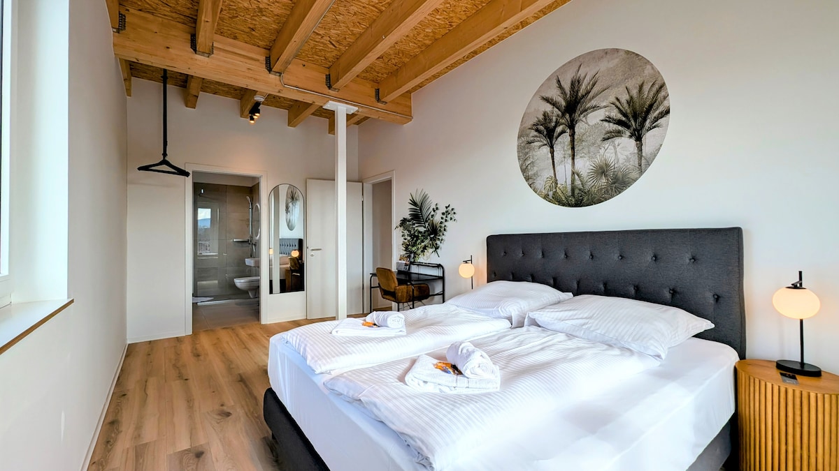 A spacious bedroom features a plush bed with soft linens and two bedside lamps. Natural light fills the room through large windows, highlighting the wooden beams on the ceiling. A tasteful round wall art displays palm trees, creating a calming focal point. A doorway leads to an adjacent bathroom.
