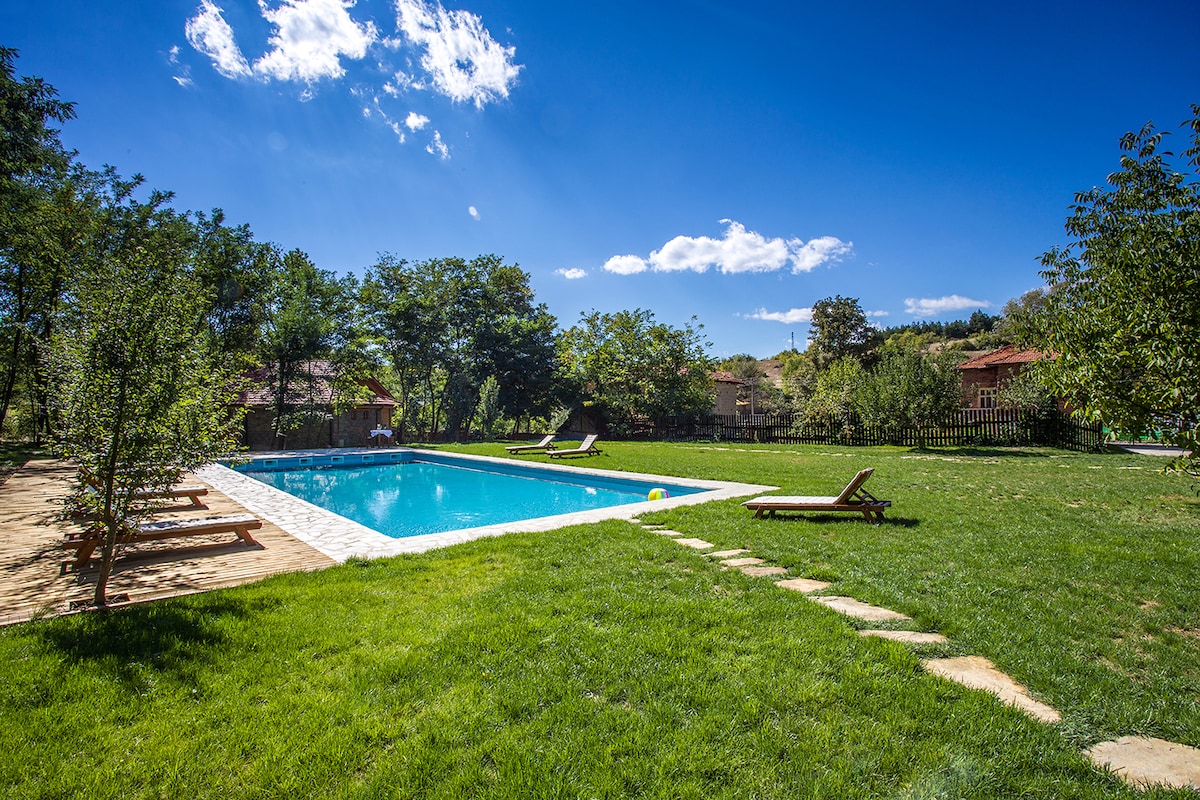A spacious outdoor area features a large swimming pool surrounded by lush green grass and trees. Sun loungers are positioned nearby for relaxation, while a pathway leads up to the pool. The clear blue sky is dotted with a few clouds, enhancing the serene setting.