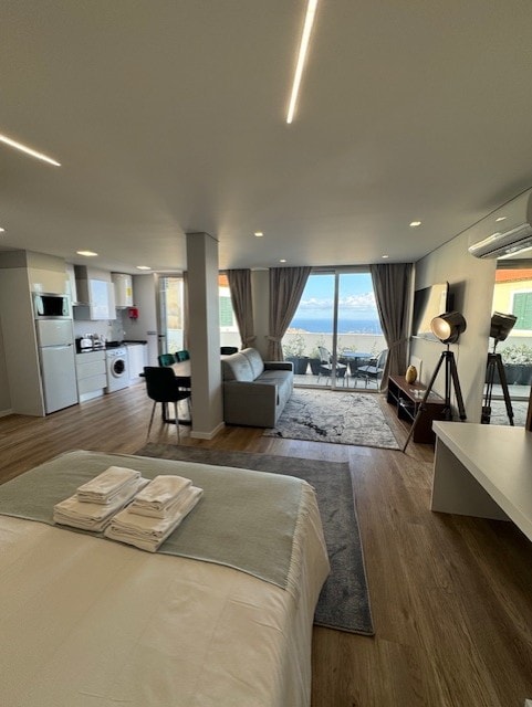 A spacious studio apartment is depicted with a cozy bed at the forefront. A kitchenette is visible, accompanied by a small dining area. Large glass doors lead to a balcony with views, allowing natural light to fill the room. Neutral color tones enhance the contemporary design.