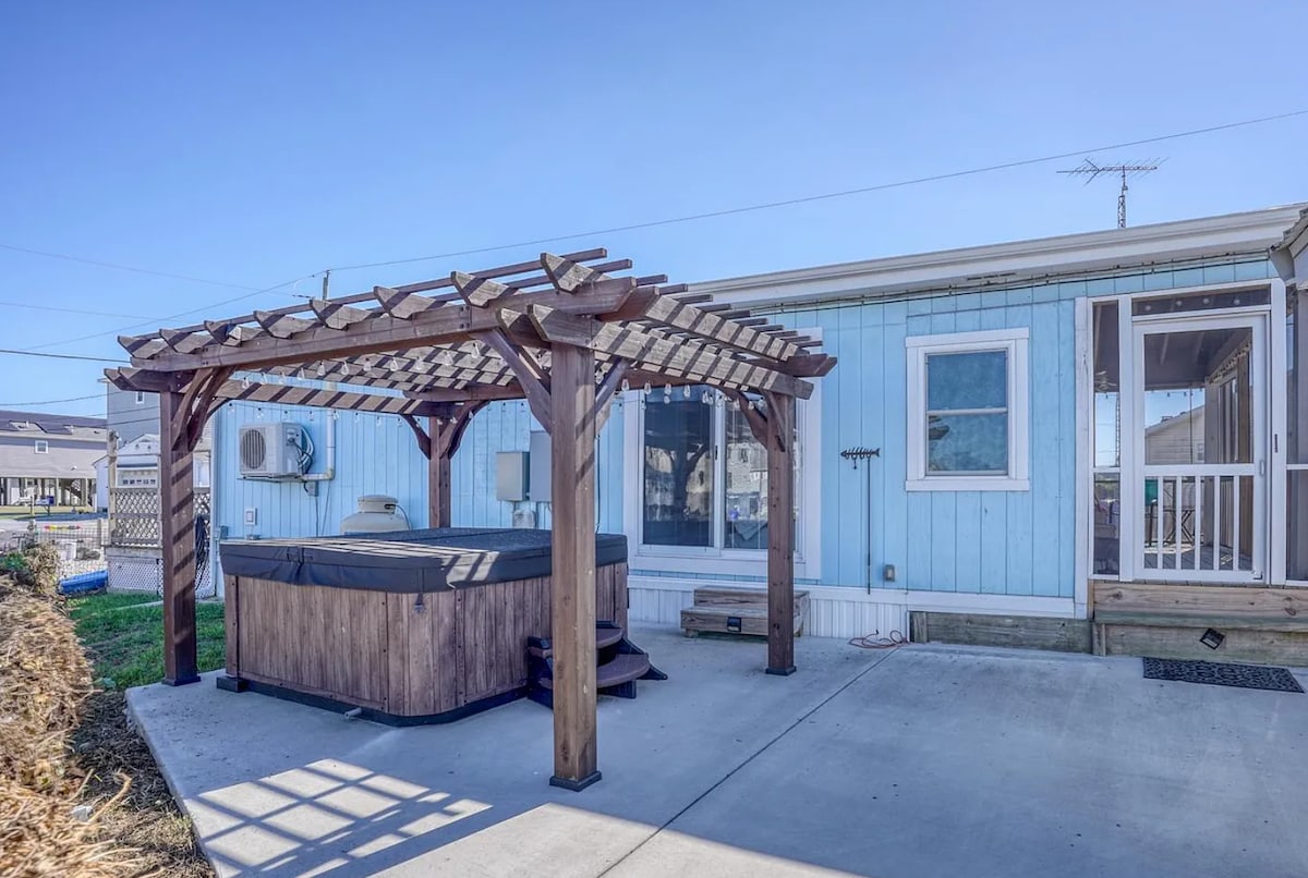 A private outdoor area showcases a hot tub beneath a wooden pergola, providing a shaded retreat. The cottage's light blue exterior complements the serene atmosphere. A concrete patio surrounds the hot tub, enhancing the outdoor space for relaxation and enjoyment.