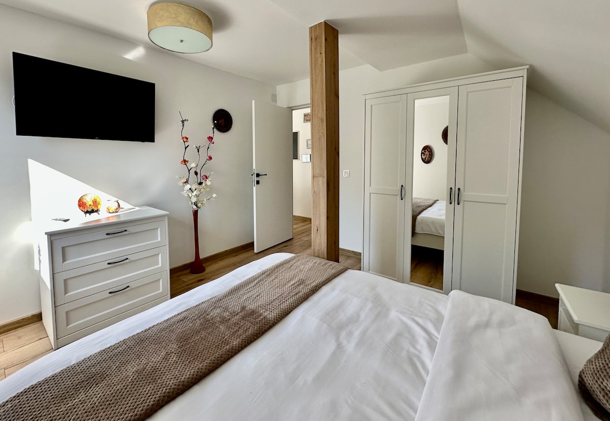 A bright and airy room features a spacious bed dressed in white linens and a neutral throw. A television is mounted on the wall, while a closet with mirrored doors reflects the light. Soft decorations, including a floral arrangement, add a subtle touch.