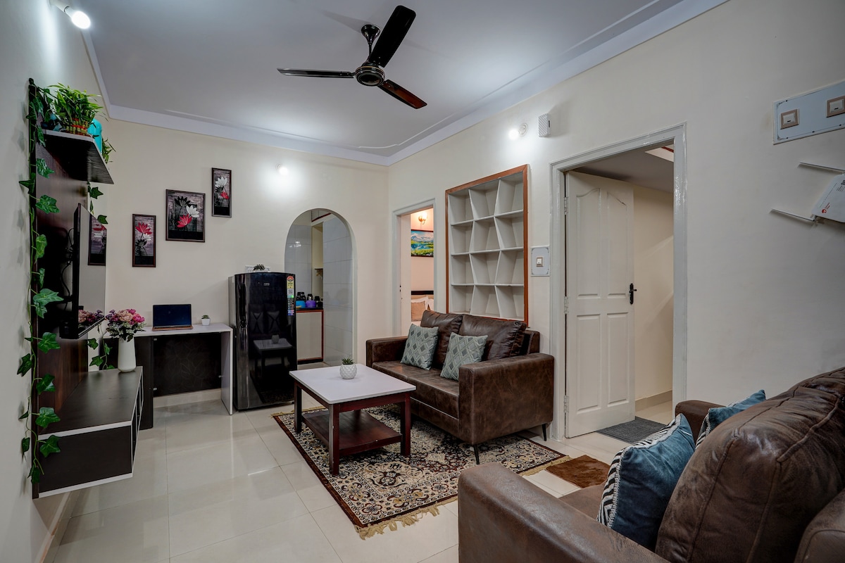 A spacious living area features a comfortable seating arrangement with plush sofas and a coffee table on a decorative rug. Wall-mounted shelves hold decorative items, while a black refrigerator is visible in the background. Natural light brightens the room through nearby windows.