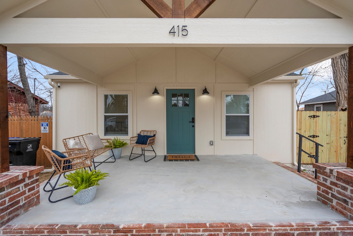 A welcoming front porch features a light blue door and two comfortable lounge chairs. Potted plants are positioned on the concrete floor, enhancing the inviting entryway. The space is sheltered by a peaked roof, providing an airy feel with natural light filtering through the nearby windows.