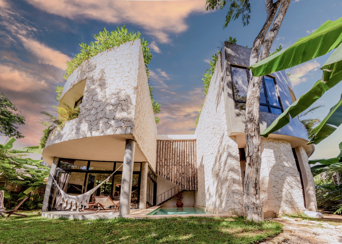 A modern villa is framed by lush greenery, featuring unique architectural elements with curved stone walls. Large glass windows provide a view of the landscaped garden and pool area, creating a harmonious blend of nature and design.