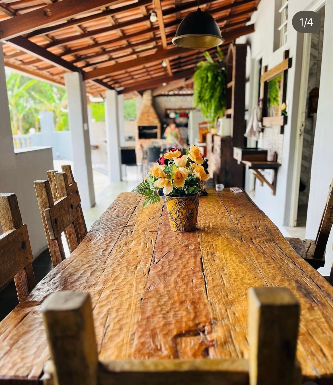 A rustic wooden dining table is set under a thatched roof, featuring a vase of flowers in the center. Surrounding chairs complement the natural wood finish, and a large open space reveals glimpses of greenery and outdoor cooking amenities in the background.