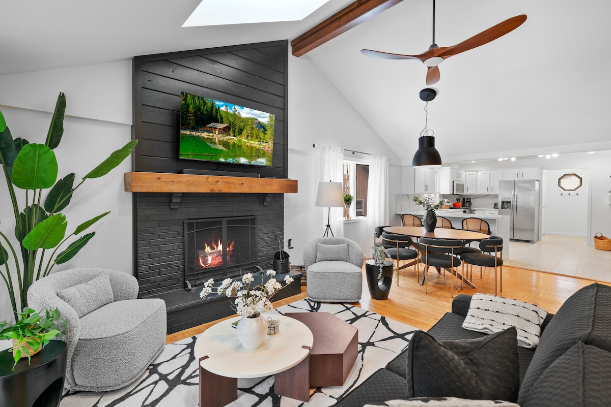 A modern living space features a black fireplace with a television mounted above it. A ceiling fan hangs overhead, and a large skylight allows natural light to fill the room. Comfortable seating arrangements include two gray armchairs and a dark sofa, while a round dining table with chairs is positioned nearby.