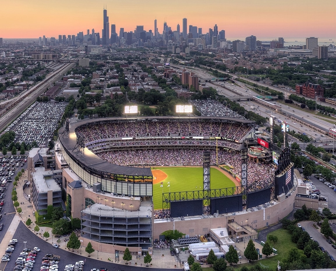 An expansive view of a baseball stadium filled with spectators is shown against the backdrop of a vibrant city skyline at sunset. The landscape includes extensive parking and distant rail lines, with illuminated stadium lights highlighting the play area.