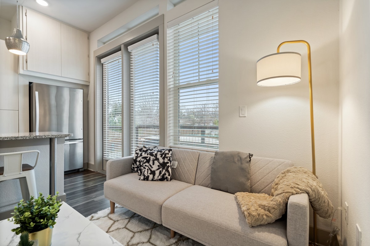 A modern living area is highlighted by a light grey sofa adorned with decorative pillows. Natural light pours in through large windows with blinds, enhancing the bright ambiance. A stylish lamp stands beside the sofa, while a small green plant adds a touch of nature.