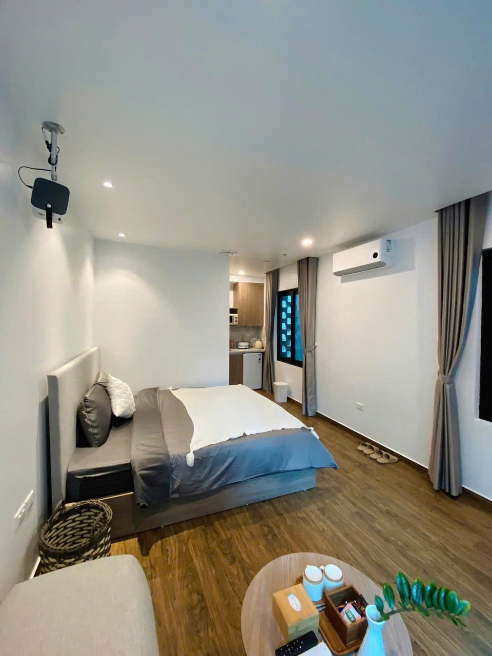 A modern studio space features a spacious bed with neutral bedding. A small kitchenette is visible in the background, equipped with essential appliances. Soft lighting highlights the warm wooden flooring, while light drapes frame a window, adding to the room's inviting ambiance.