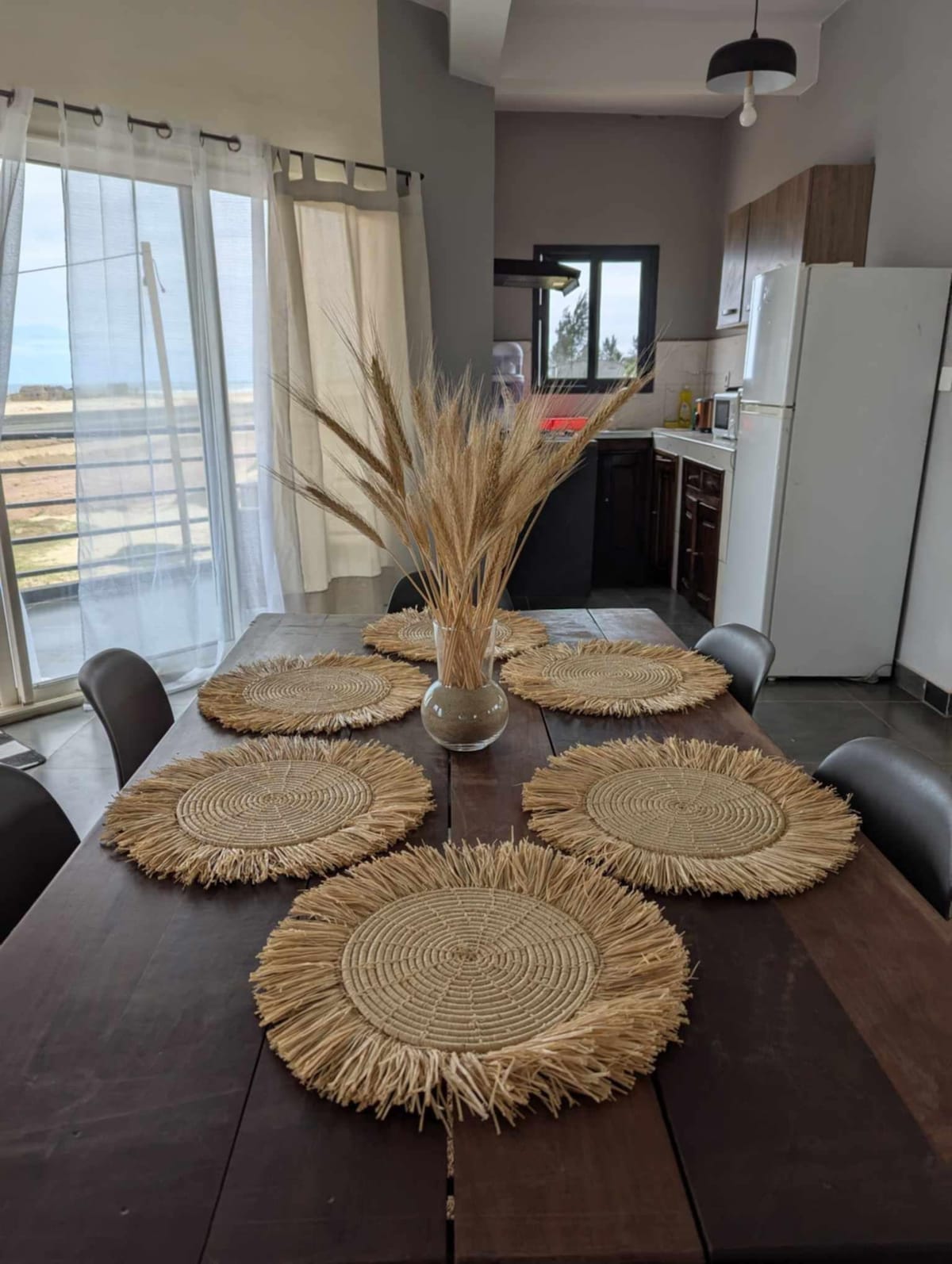 A dining table is set with six round placemats made from natural fibers. A vase filled with dried grass stands centrally. The kitchen area is visible in the background, featuring modern cabinetry and a refrigerator, with natural light entering through large windows.