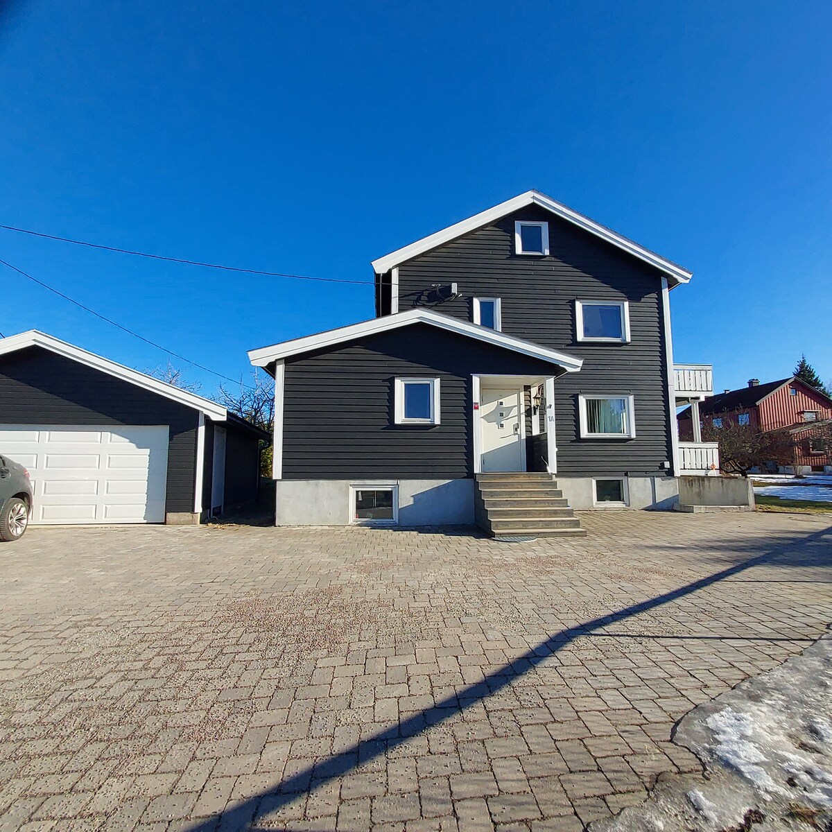 The exterior of a three-story building is shown, featuring dark wood siding and a sloped roof. A set of steps leads up to the entrance, with a paved driveway and garage visible beside the structure. Clear blue skies provide a bright backdrop.