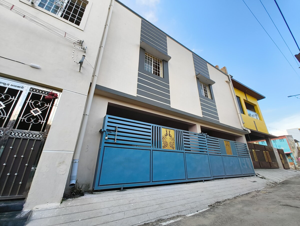 The exterior of a modern building is presented, featuring a sleek blue gate with decorative designs. The walls are painted in a light color, and multiple windows are visible on the upper level. The surroundings are urban, with additional buildings seen in the background.