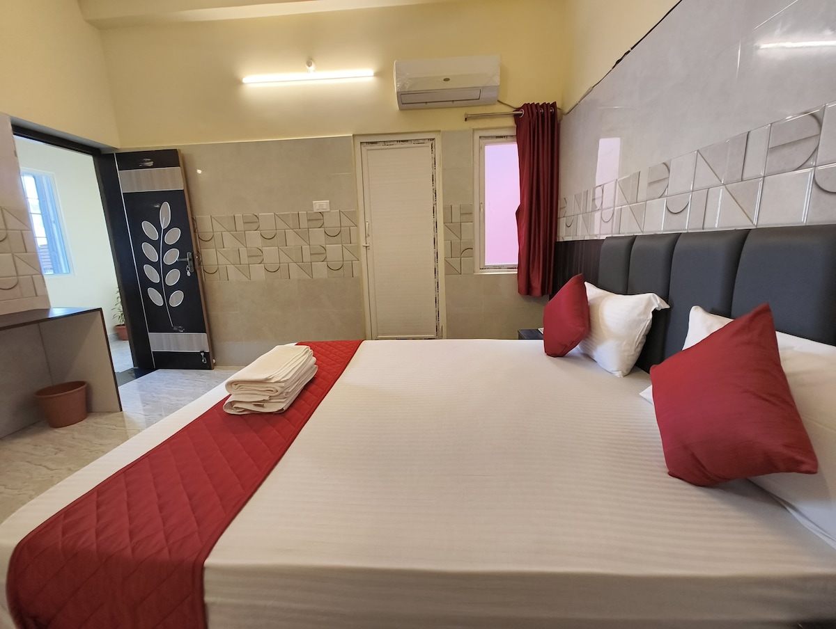 A spacious bedroom is shown, featuring a large bed with a red and white color scheme. Two red pillows are placed at the head of the bed, and neatly folded towels are positioned on the foot of the bed. A window with partially closed curtains is visible, allowing soft light to enter the room.