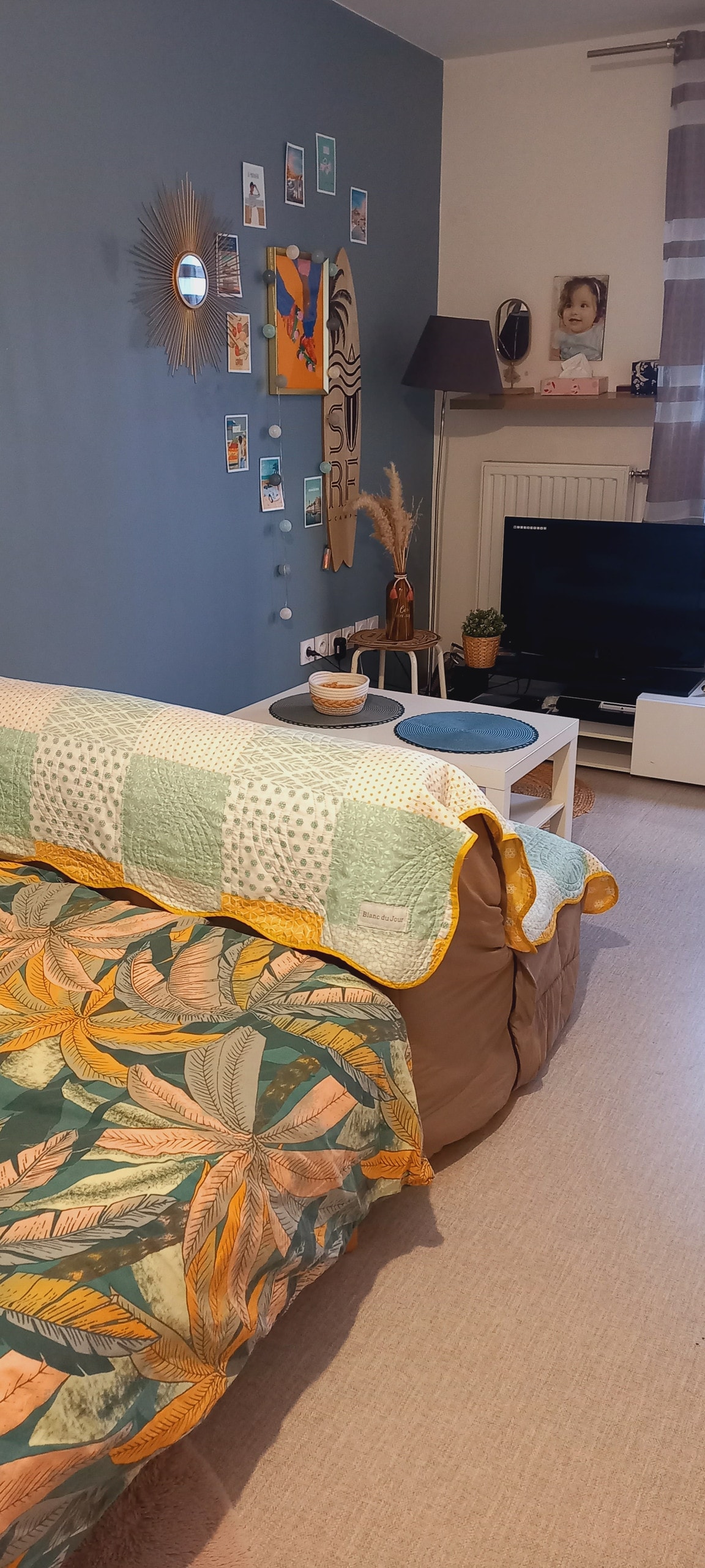 A well-appointed studio features a cozy sleeping area with a patterned blanket covering the bed. A gallery wall is adorned with photography, and a small coffee table is positioned in front of a TV. Natural light filters through window treatments, creating a welcoming ambience.