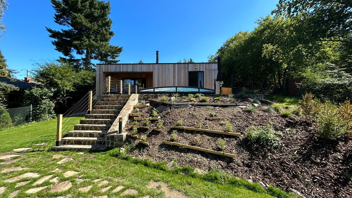 A modern structure is set against a clear blue sky, featuring a wooden façade and large glass doors. Stone steps lead to a landscaped area with tiered garden beds and greenery, creating a natural transition between the home and surrounding nature.
