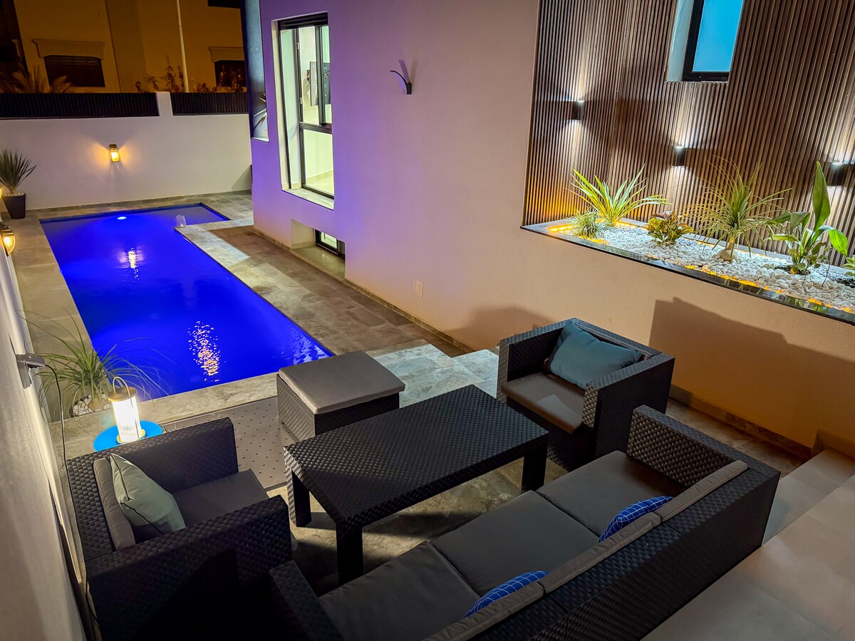 An outdoor seating area features dark wicker furniture arranged around a black table, positioned near a rectangular pool illuminated in blue. A modern stone wall and potted plants enhance the serene ambiance. Soft lighting creates a calming atmosphere, inviting relaxation in the evening.