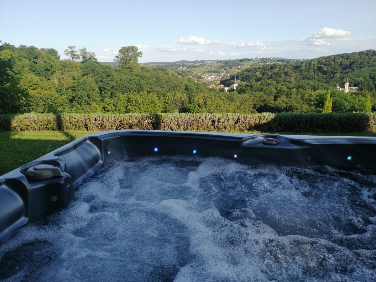 A hot tub filled with bubbling water is situated in the foreground, providing a view of rolling green hills in the background. The landscape is dotted with trees and distant buildings, revealing a serene natural environment under a clear blue sky.