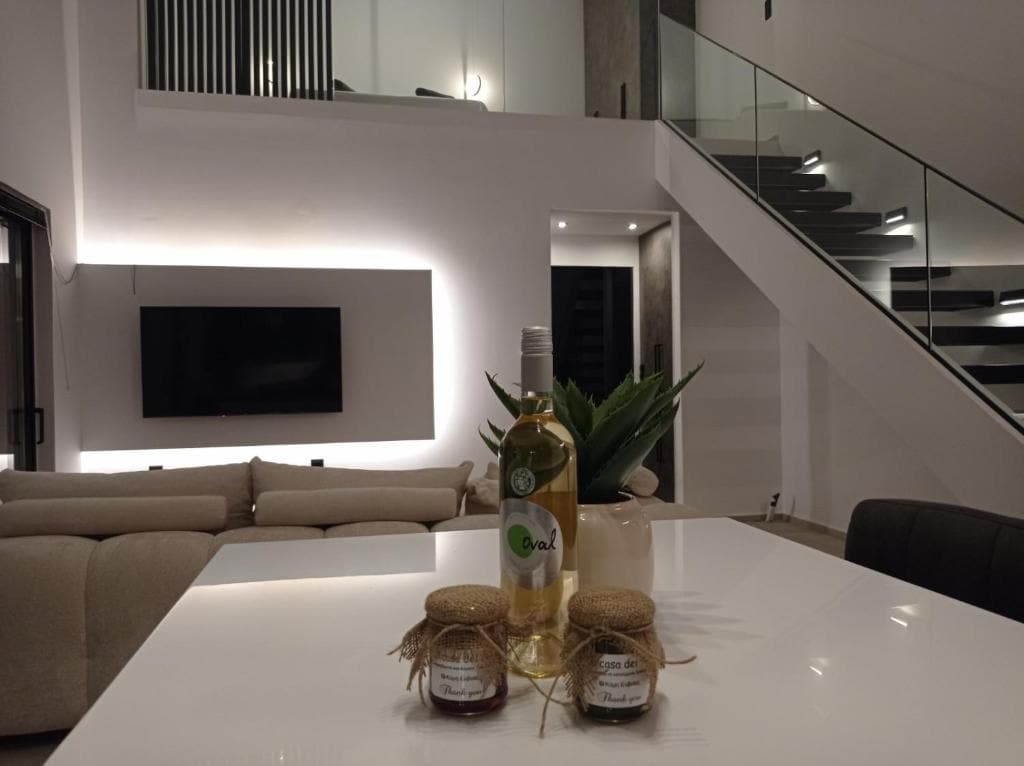 A modern living area is highlighted, featuring a large, light-colored sofa and a wall-mounted television. A white countertop displays two jars with decorative lids and a bottle of wine, alongside a green plant centerpiece. Soft lighting creates a welcoming ambiance.