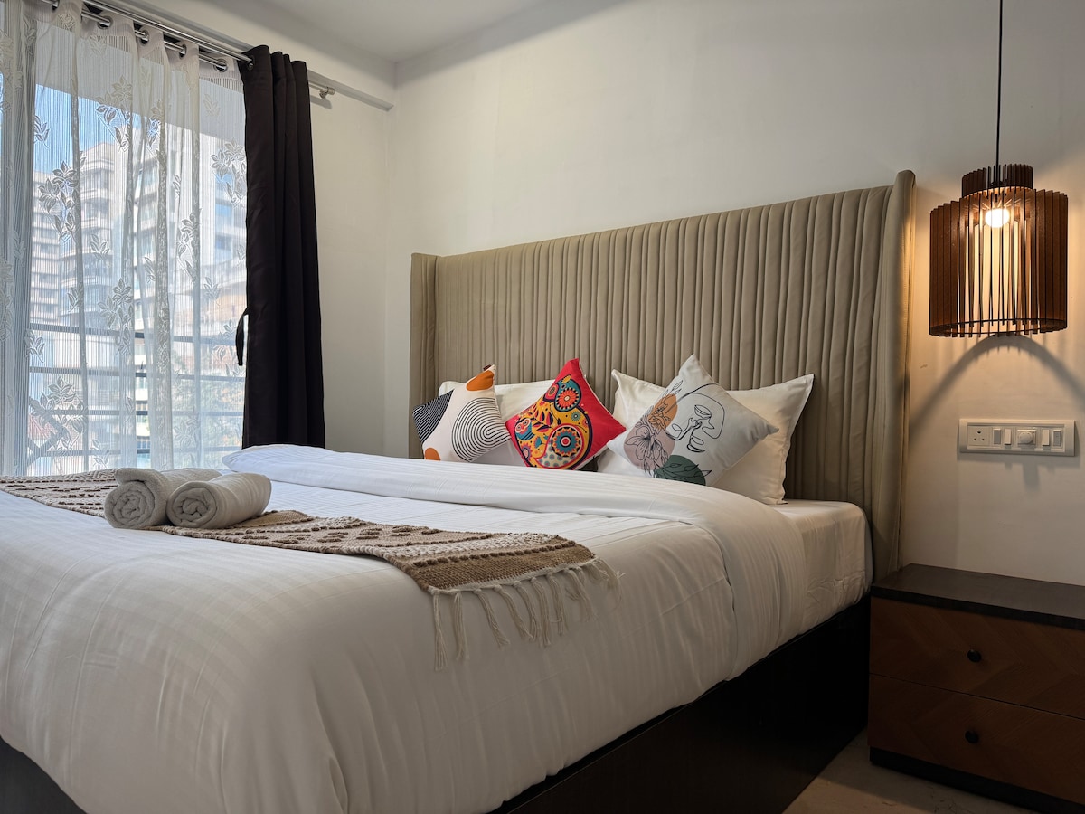 A comfortable king-size bed is displayed, adorned with a cozy comforter and decorative pillows. Natural light filters through a large window, complemented by sheer curtains. A bedside lamp casts a soft glow, enhancing the calm ambiance of the room.