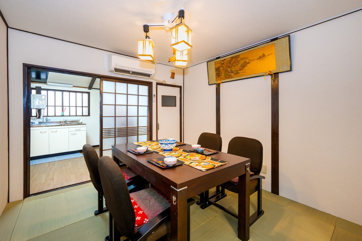 A traditional dining area is presented with a wooden table surrounded by four chairs. A variety of dishes is displayed on the table, with soft lighting illuminating the space. The tatami flooring adds to the authentic Japanese ambiance, visible through sliding doors leading to the kitchen.
