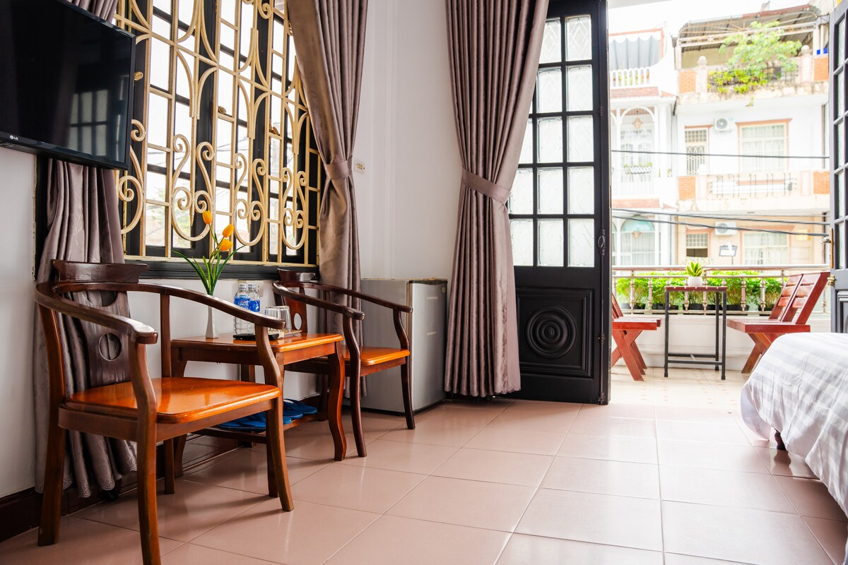 A small seating area is positioned near a large window, featuring two wooden chairs and a small table. The space is illuminated by natural light and decorated with light curtains. A mini-fridge and complimentary water bottles are visible in the corner.