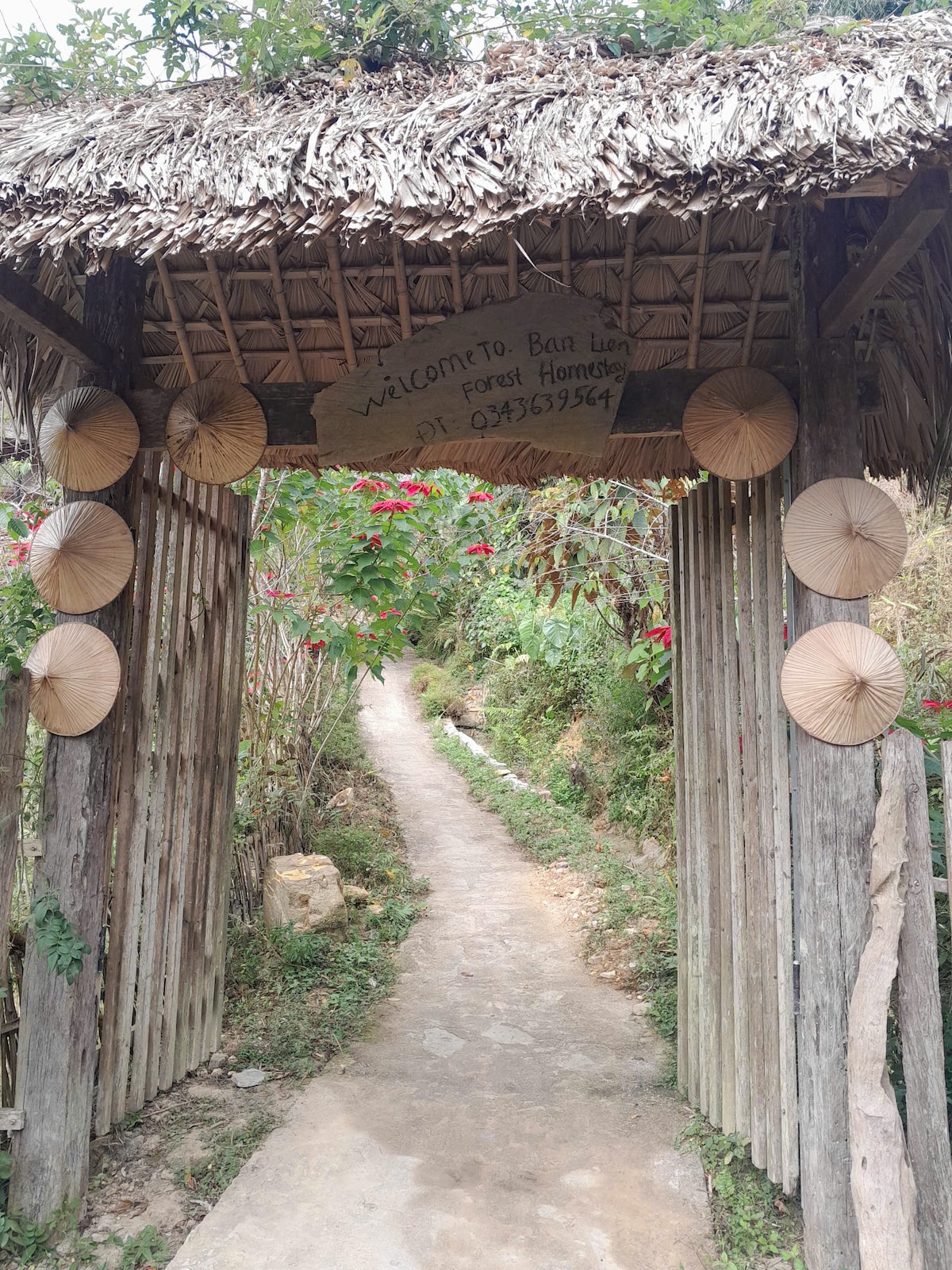 An inviting entrance is framed by a thatched roof, featuring hand-painted signage and decorative fans. A stone path winds through lush greenery, leading to the property, while colorful flowers enhance the natural setting.