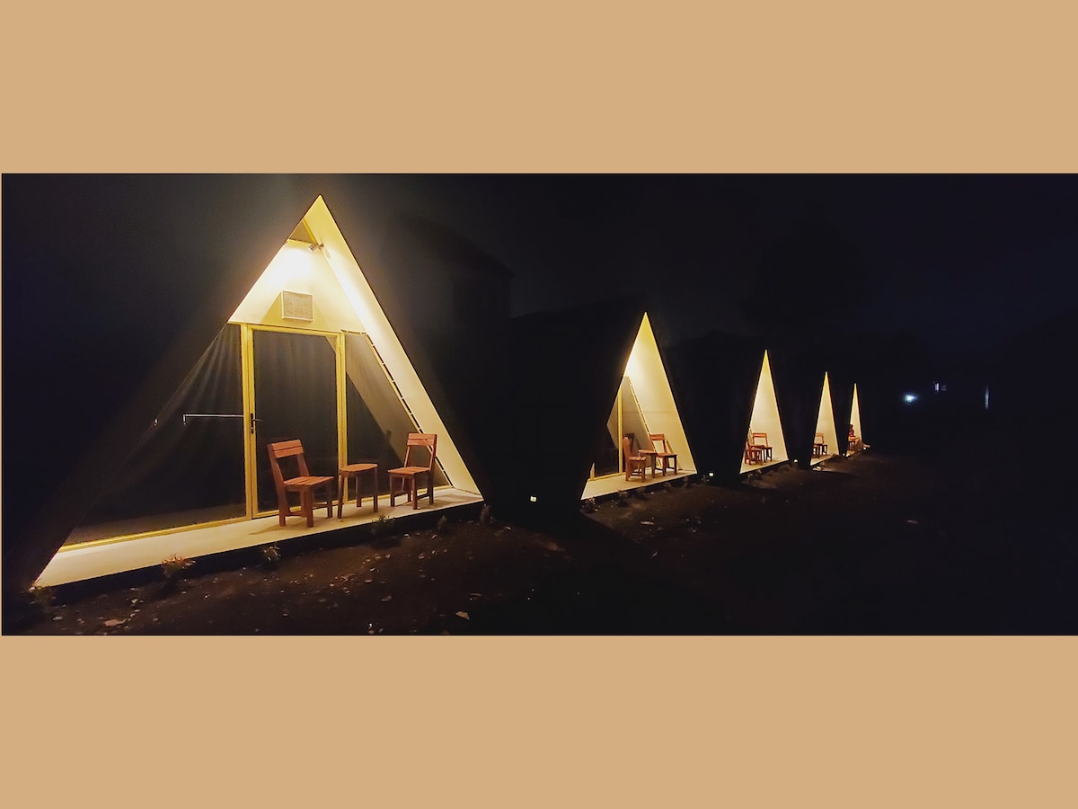 Four triangular cabins are illuminated softly from within, each featuring a pair of wooden chairs positioned outside. The calm night scene highlights the architectural design and extends into the surrounding darkness, creating a serene and inviting ambiance.