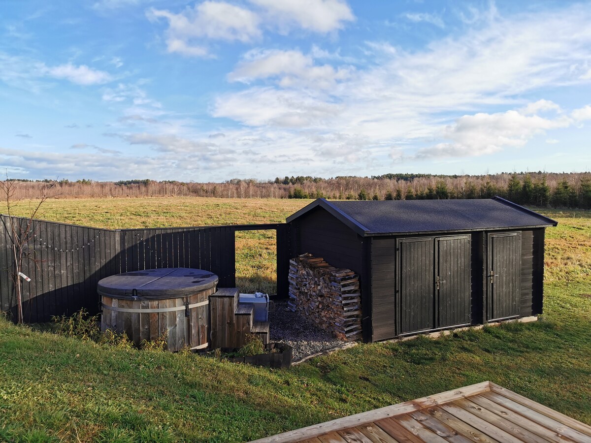 A view captures a wooden hot tub and a black wooden shed, both set against a backdrop of sprawling fields and a partly cloudy sky. The hot tub is surrounded by a wooden deck, while a stack of firewood leans beside the shed.