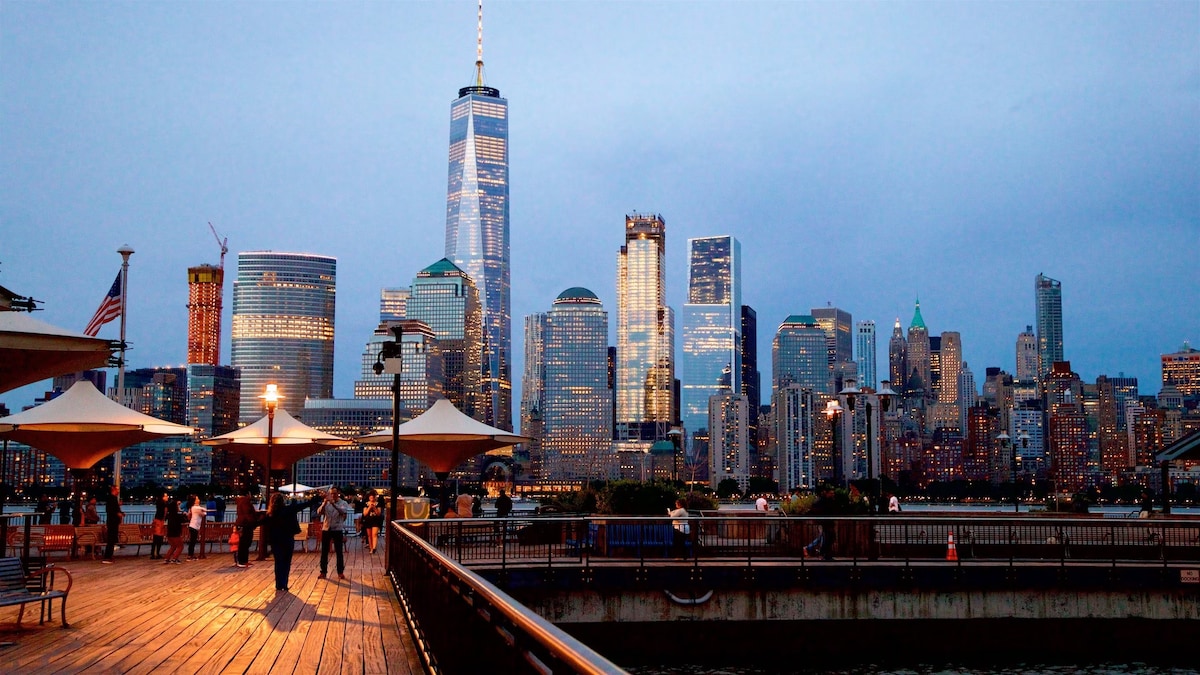 The New York City skyline is displayed at dusk, showcasing illuminated skyscrapers including One World Trade Center. A waterfront boardwalk is visible in the foreground, where visitors can be seen enjoying the view under the open sky. Soft lighting from lamps adds to the atmosphere.