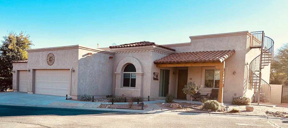 The exterior of a single-story home is featured, showcasing a stucco facade with a tile roof and arched windows. A two-car garage is visible, along with a spiral staircase leading to a second-floor space. Desert landscaping enhances the charming curb appeal.