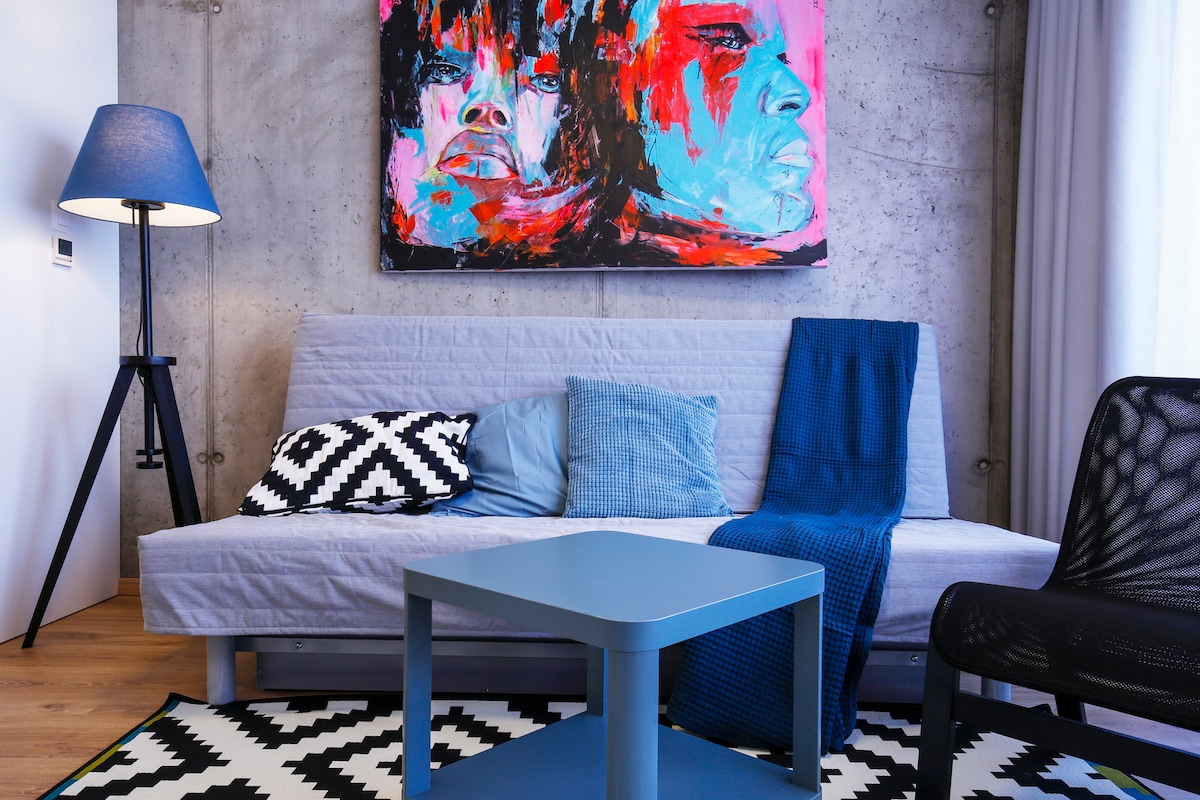 A modern living area features a gray sofa adorned with patterned cushions, complemented by a solid blue throw. A blue coffee table is situated in front of the sofa, resting on a monochrome geometric rug. A standing lamp provides soft illumination beside the seating area.