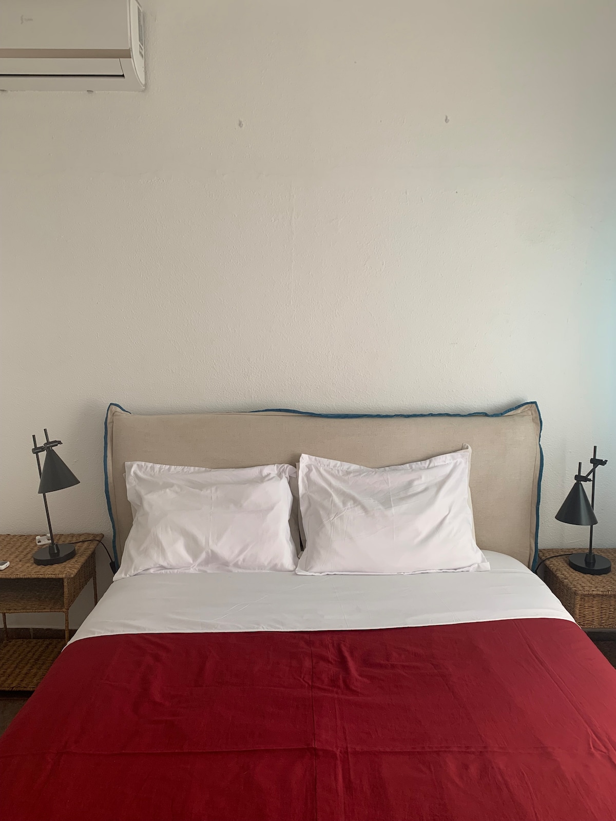 A cozy sleeping area features a spacious bed with a neutral headboard and a red throw covering the foot. Two nightstands with modern lamps are positioned on either side of the bed, complemented by light streaming in from a window.
