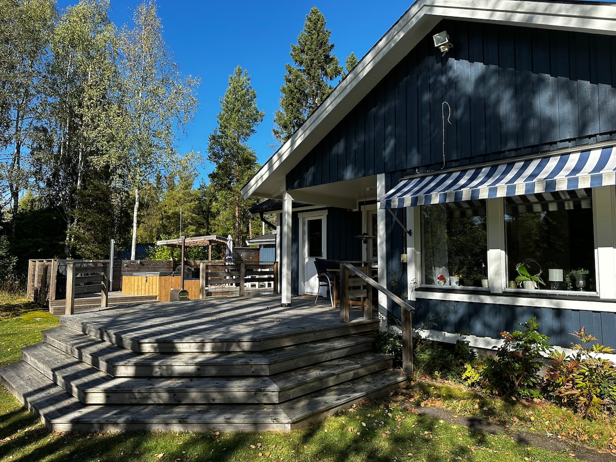 A spacious outdoor deck features wooden steps leading up to the entrance of a blue house. Large windows with striped awnings allow for ample natural light. A relaxing hot tub and garden area are visible in the background, surrounded by trees and greenery.